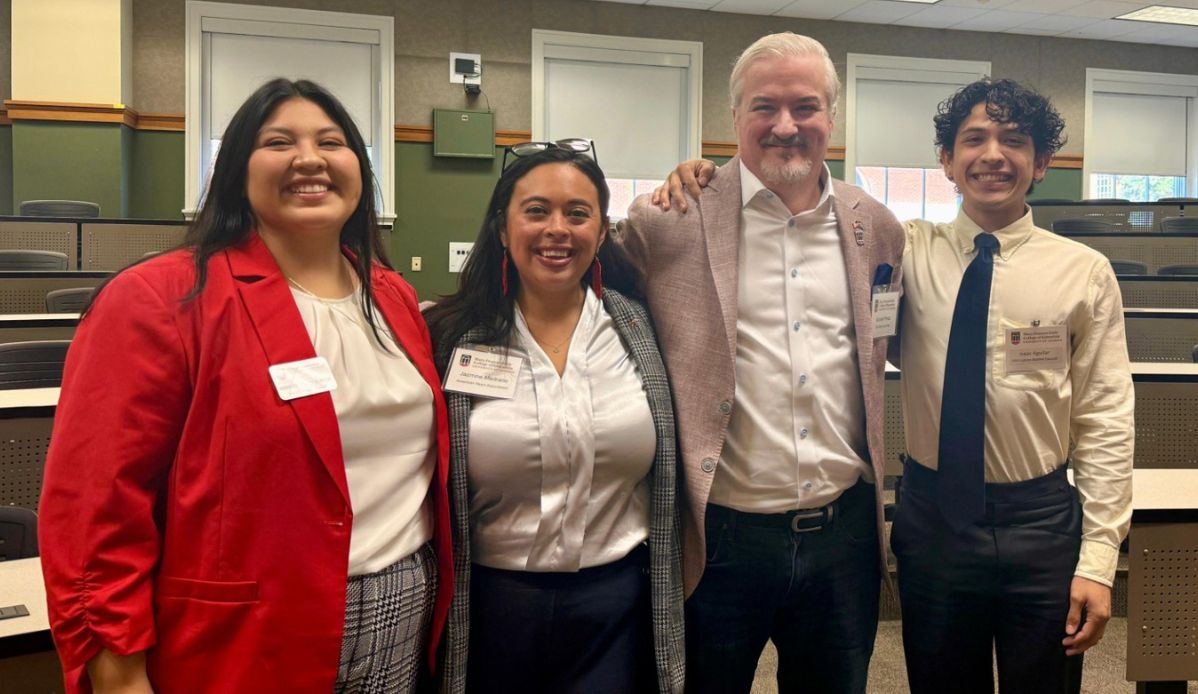 Four Latino Alumni Council Members smile together at an event in a classroom.