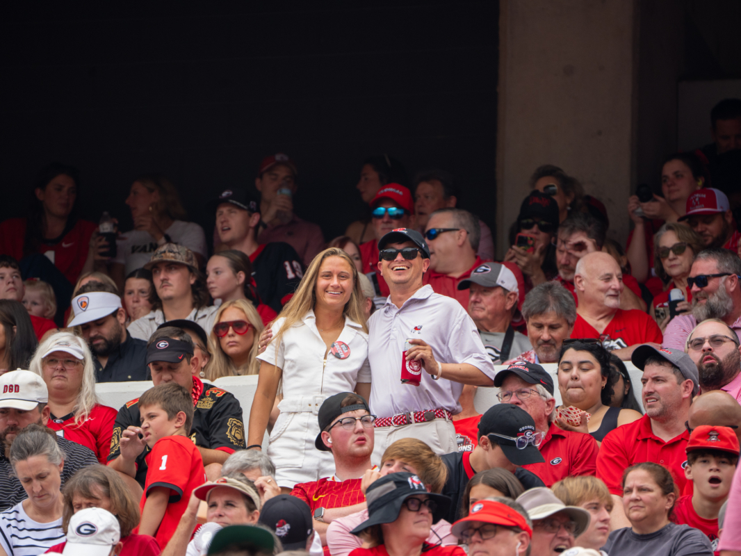 A man and a woman are standing up in a crowd of UGA fans. The women wears white and the man wears khakis and a button up shirt and holds a beverage.