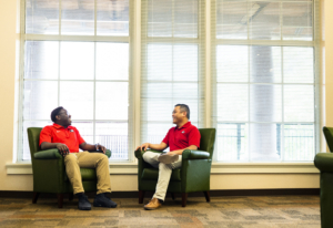 Two people in red UGA polos sit in green armchairs by tall windows, laughing together